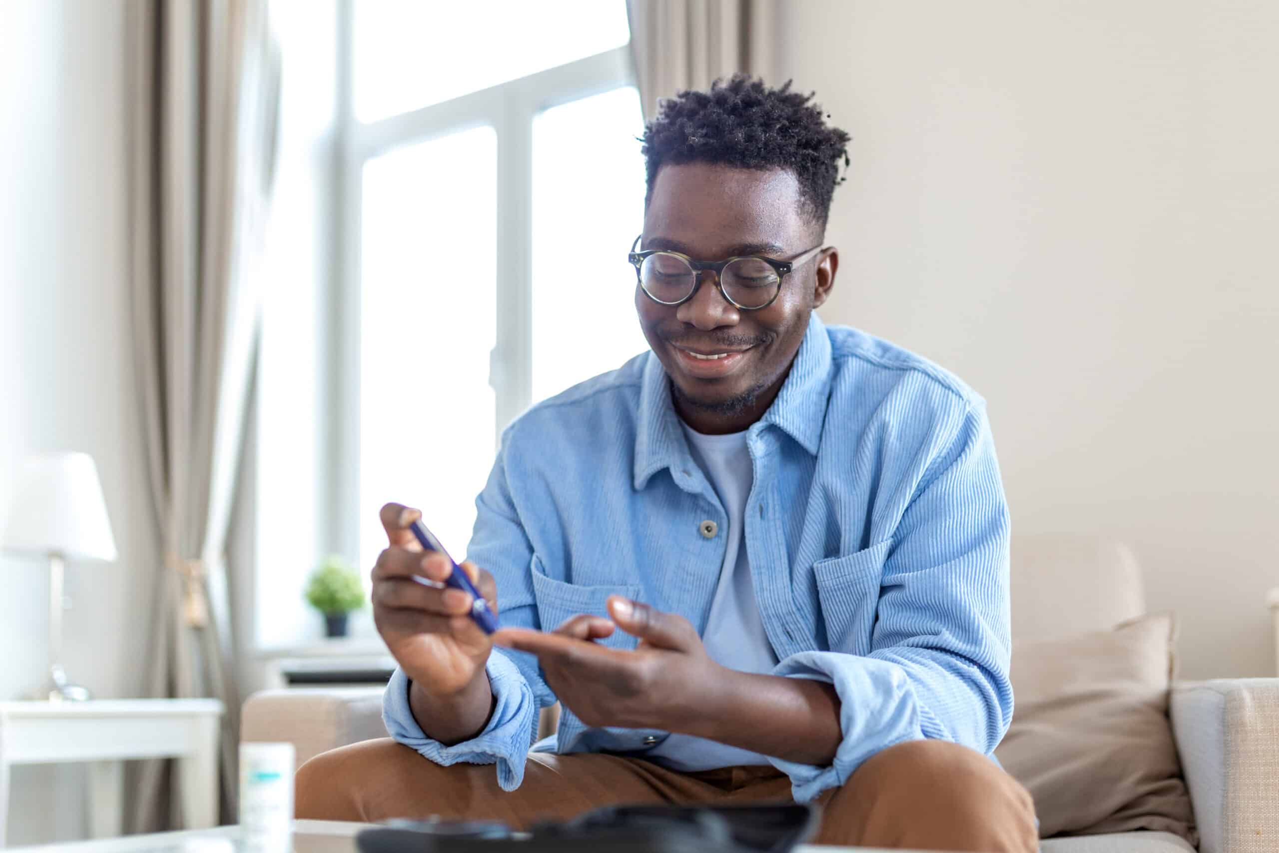 African man is sitting at the sofa at the home and taking blood from his finger due to diabetes. The daily life of a man of African-American ethnicity person with a chronic illness who is using glucose tester.