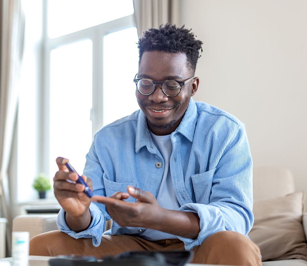 African man is sitting at the sofa at the home and taking blood from his finger due to diabetes. The daily life of a man of African-American ethnicity person with a chronic illness who is using glucose tester. T1D testing blood sugar